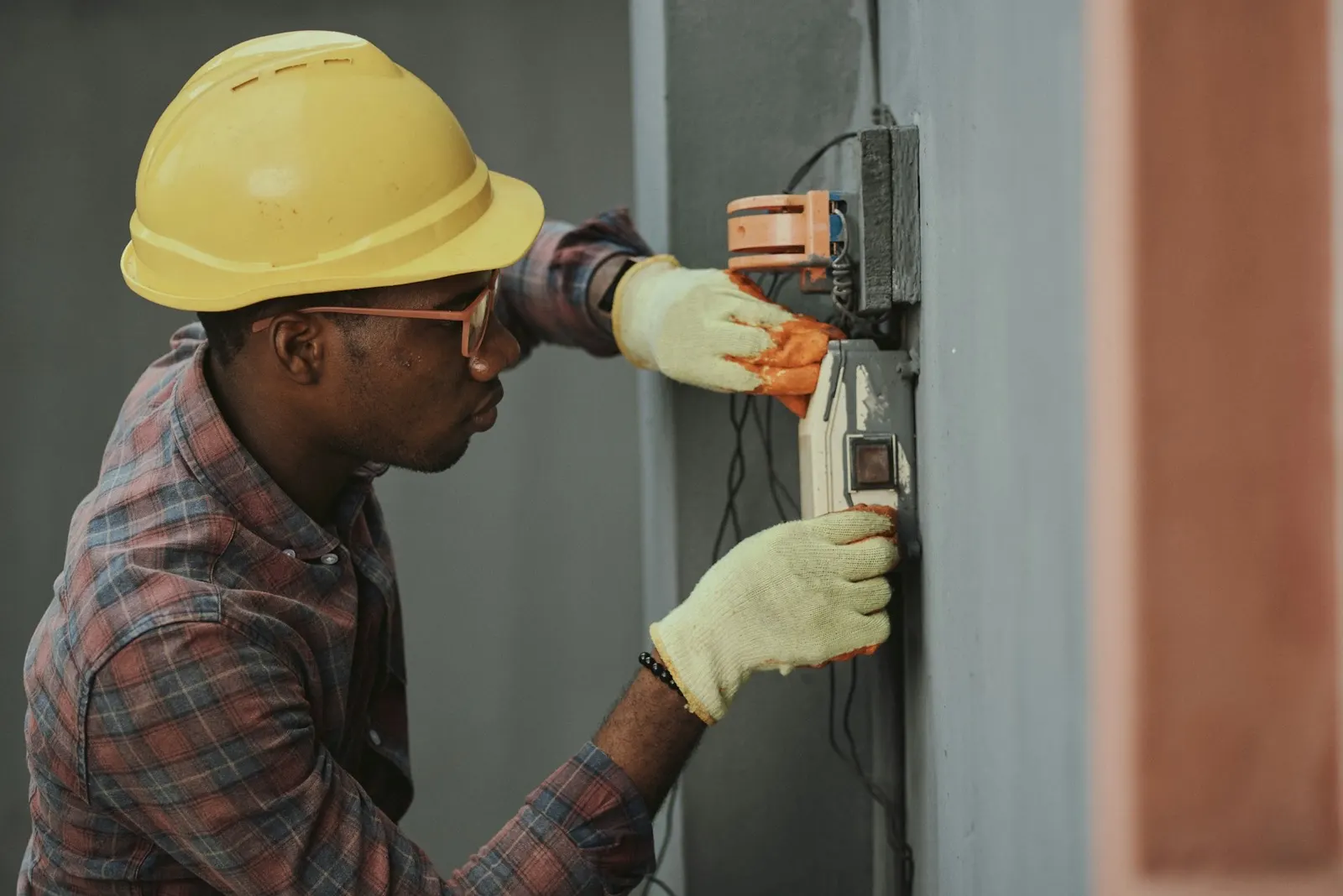 Electrician in a yellow hard hat and work gloves wiring a wall-mounted electrical box