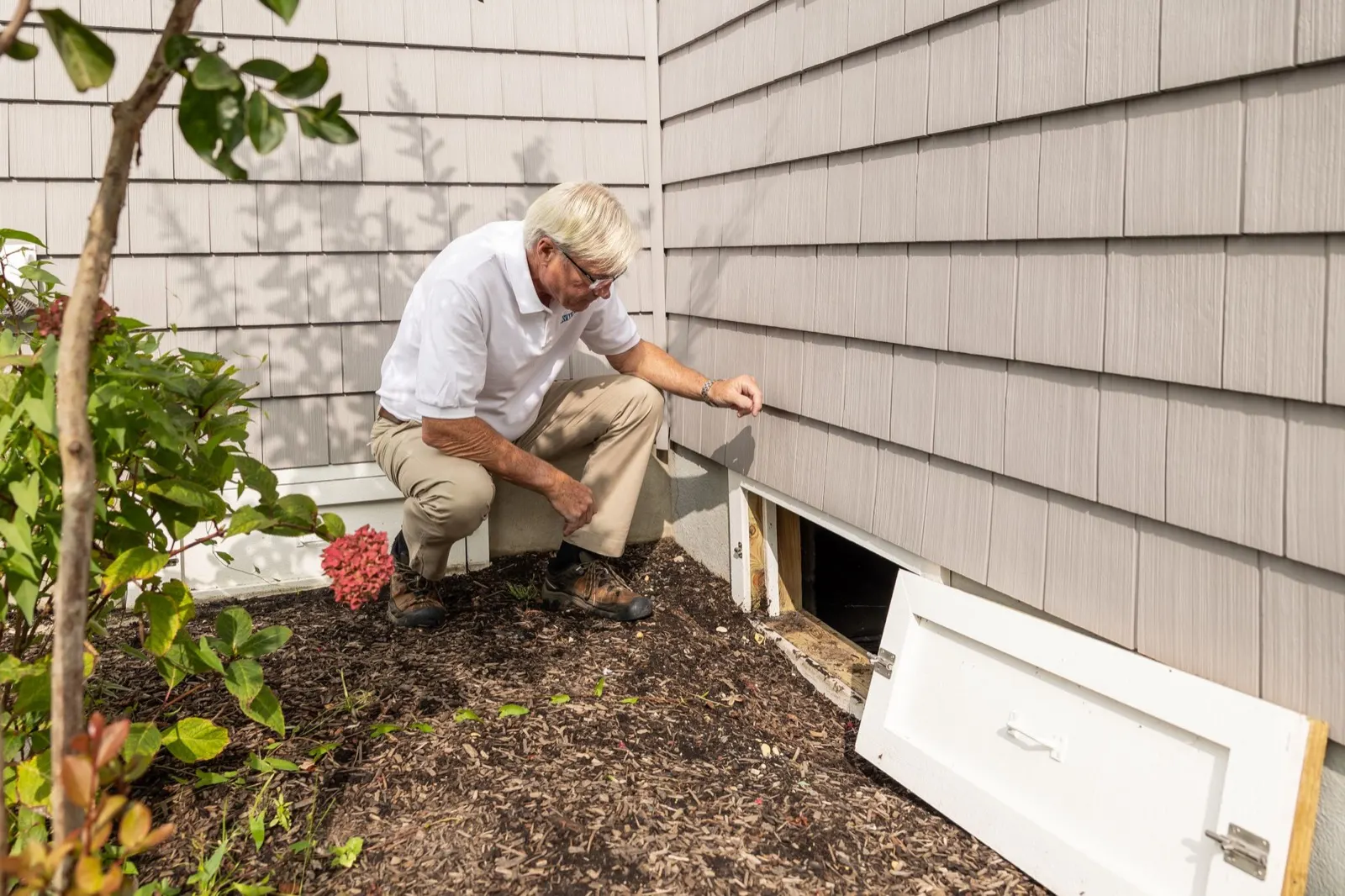 Inspector crouching beside an open crawl space vent on a house exterior