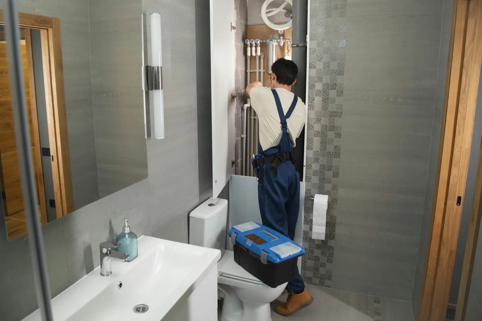 Plumber in overalls working on bathroom pipes with a toolbox on the toilet