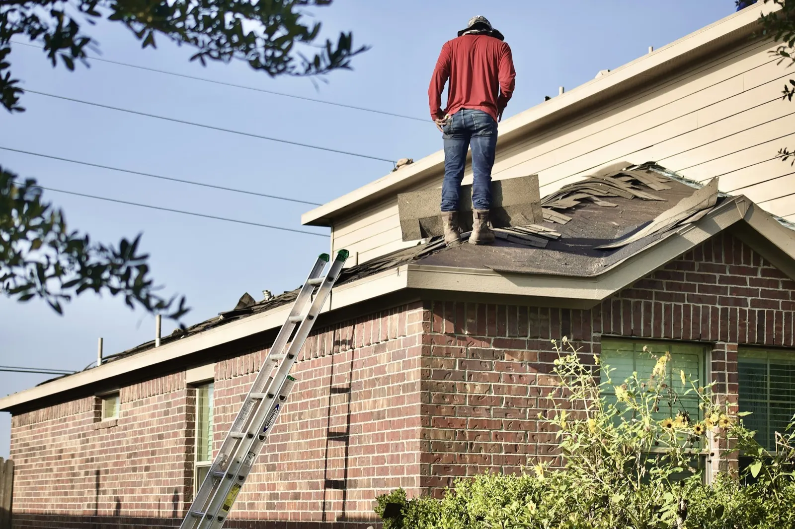 Roofer standing on a partly stripped shingle roof above a brick house with a ladder